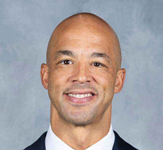 Headshot of a bald man smiling, wearing a suit and tie against a neutral background.