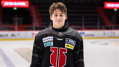 A young male hockey player wearing a black jersey with various sponsorship logos, standing on an ice rink.