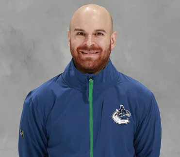 A smiling man with a bald head and beard, wearing a blue jacket with the logo of the Vancouver Canucks.
