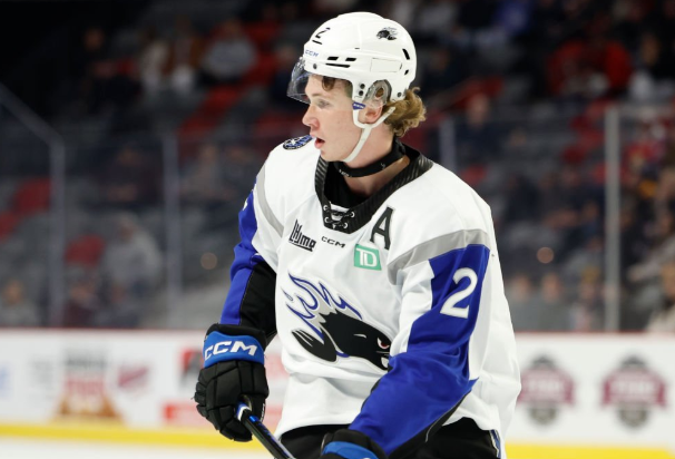 A young hockey player wearing a white jersey with blue accents and the number 2, skating on the ice during a game, with a focused expression and short, curly hair.