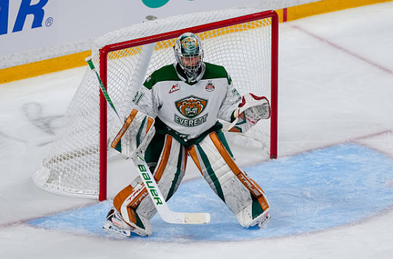 A hockey goalie in a green and white uniform standing in front of the net, preparing to block a shot. The ice rink is visible in the background.
