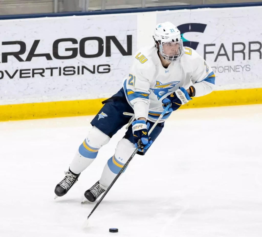 A hockey player wearing a white and blue jersey gliding on ice while controlling a puck with a stick during a game.