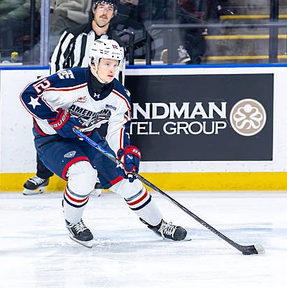 A young ice hockey player in a blue and white uniform skating on the rink while handling a hockey stick. A referee is visible in the background, and there is a sponsorship board for a hotel group.
