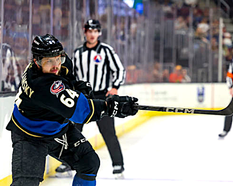 A hockey player in a black jersey with a blue stripe takes a shot on the ice, with a referee visible in the background.